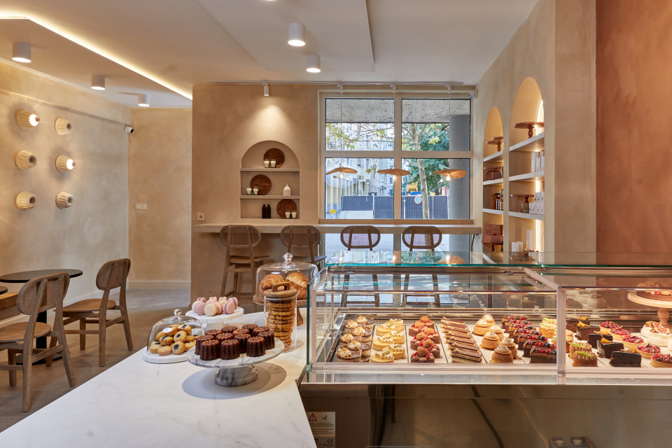 Interior of a modern pastry shop with dessert display cases, a counter with a Topciment® microcement countertop, a bar with stools, and arched shelving.
